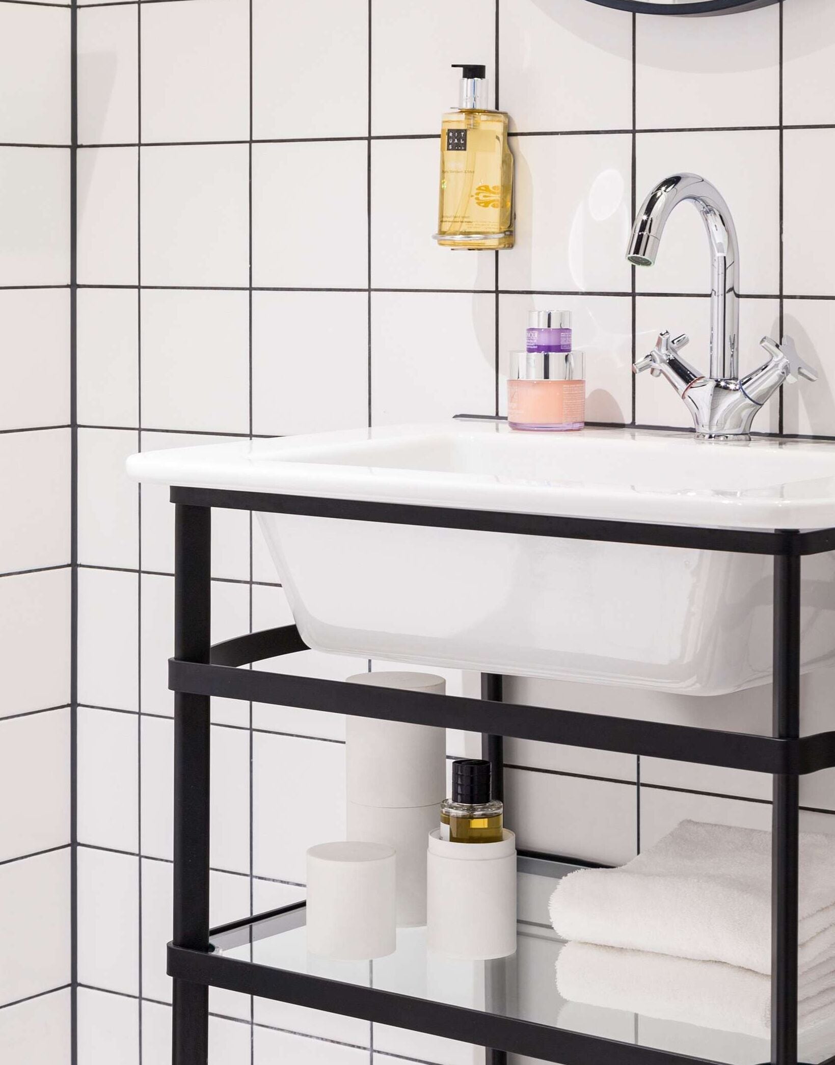 A bathroom with a white sink, black metal frame, and a mirror above. There are toiletries on the sink and shelves below.