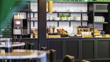 A modern cafe with a green-tiled backsplash, a coffee machine, and a variety of food and drink items displayed on shelves.