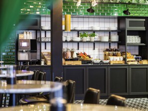 Een modern café met een groene betegelde achterwand, een koffieapparaat en diverse etens- en drankproducten die op planken staan uitgestald.