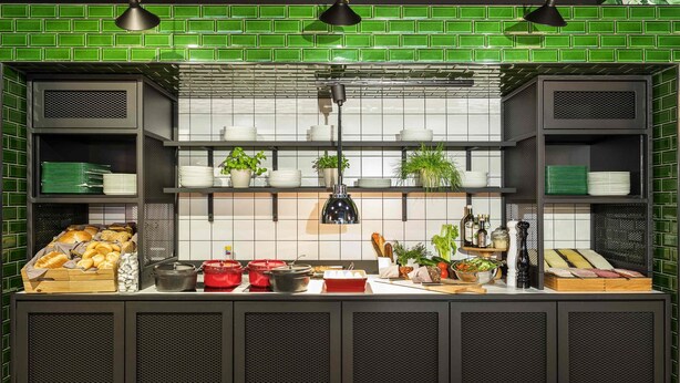Een keuken met een groene betegelde spatwand, planken met borden en kommen, en diverse etenswaren.