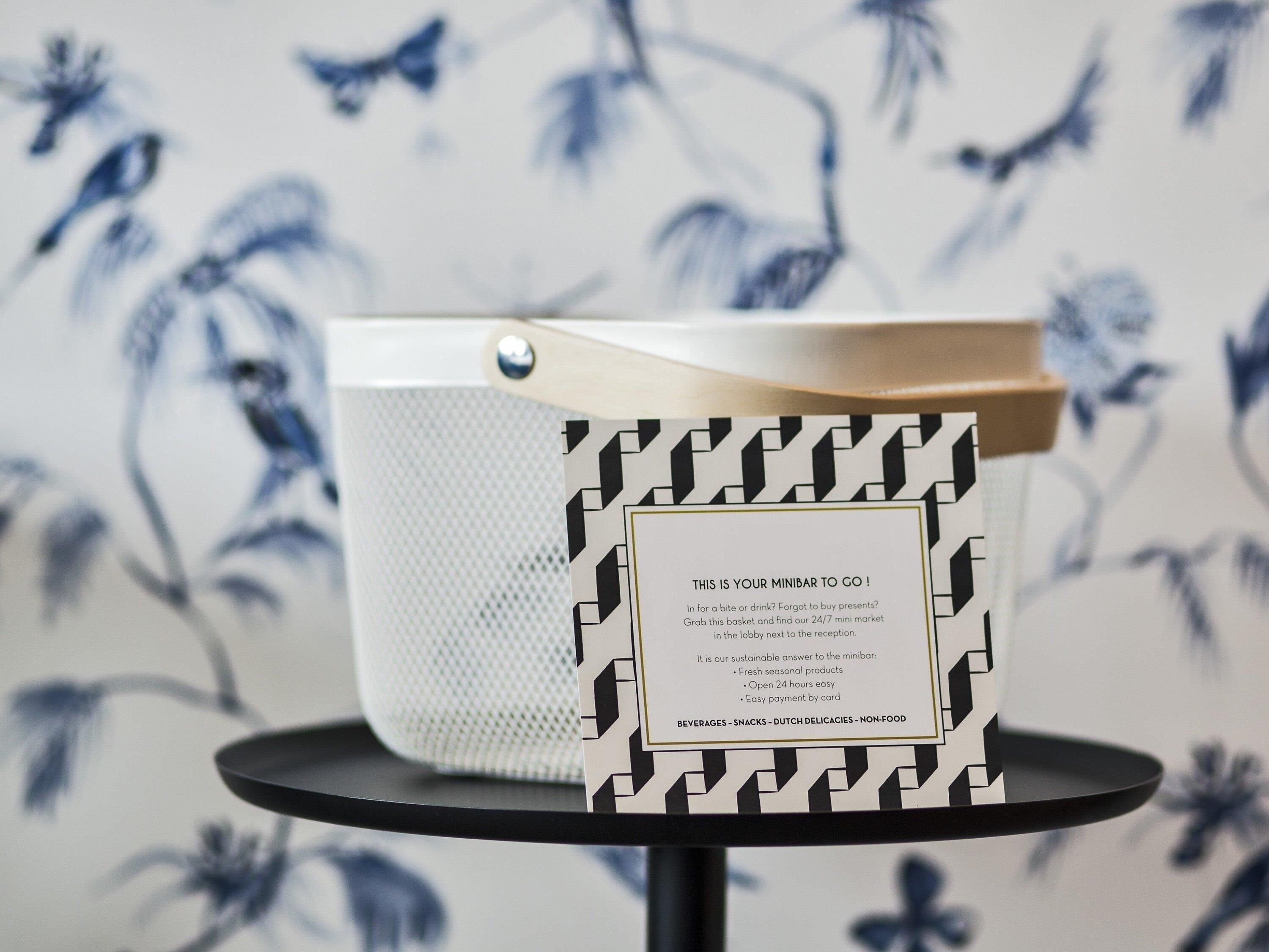 A black table with a white basket containing a sign. The sign reads 'THIS IS YOUR MINIBAR TO GO!' and lists items like beverages, snacks, and Dutch delicacies.