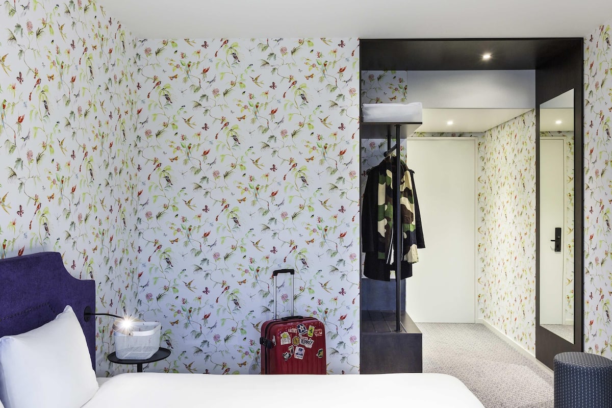 Une chambre d’hôtel avec un papier peint à motifs floraux, un lit, une valise, un porte-manteau et un miroir.
