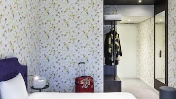 Habitación de hotel con papel tapiz floral, una cama, una maleta, un perchero y un espejo.