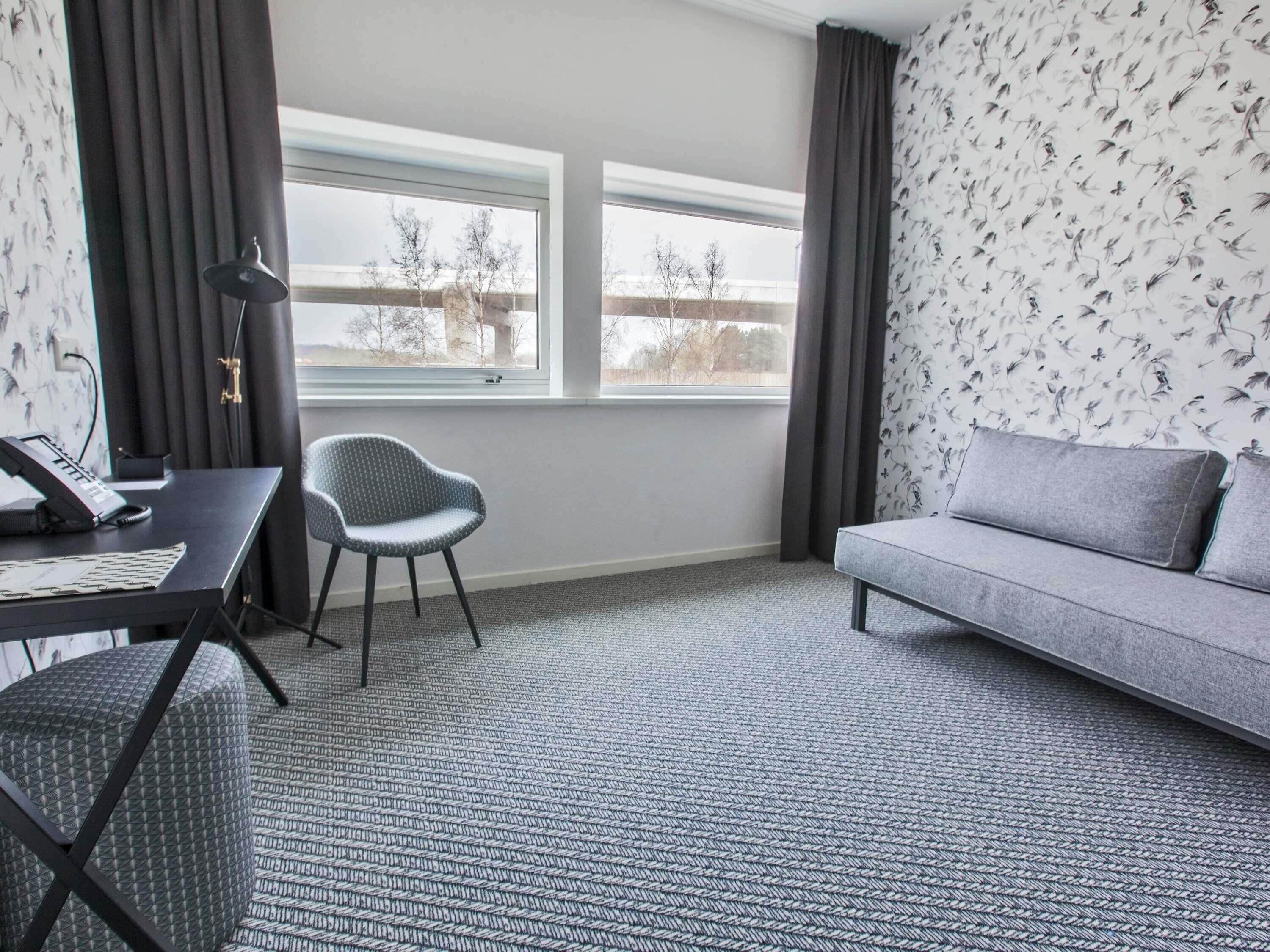 Una habitación con escritorio, silla, sofá y dos ventanas.