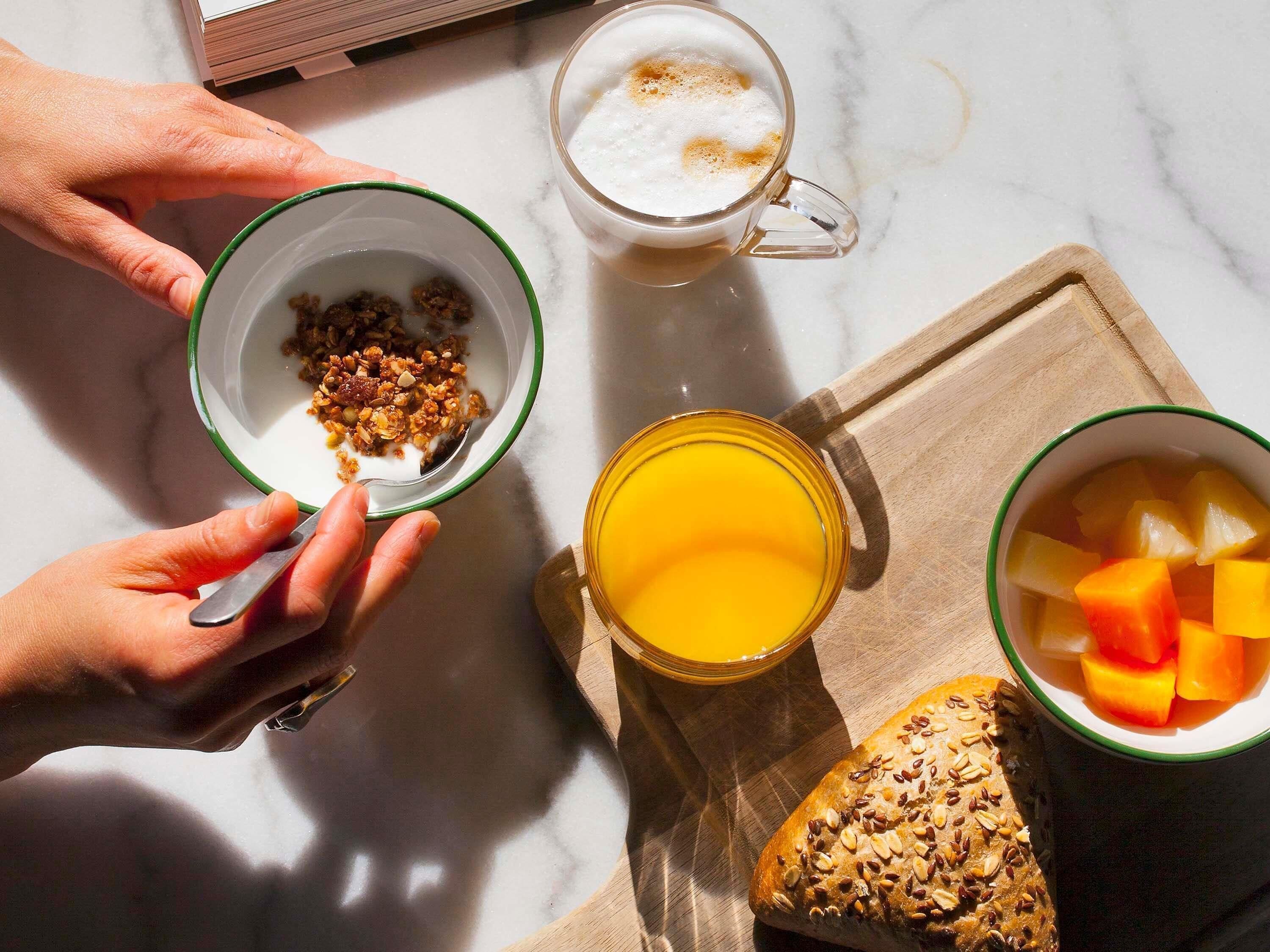 Disposição para o café da manhã com uma tigela de granola, uma caneca de café, um copo de suco de laranja e uma fatia de pão sobre uma superfície de mármore.