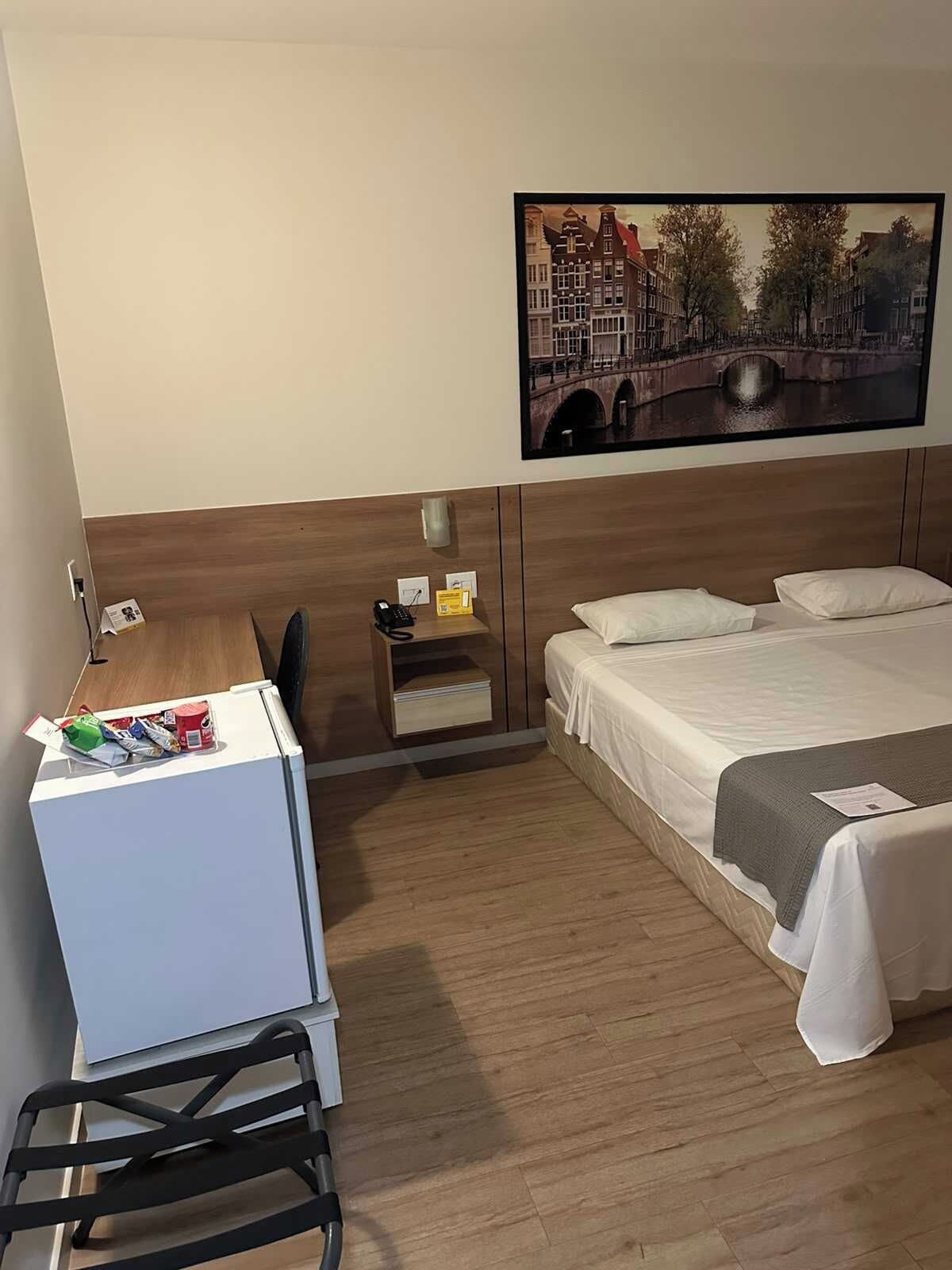 Quarto de hotel com cama, escrivaninha, cadeira, mesinha e pintura de uma paisagem urbana.
