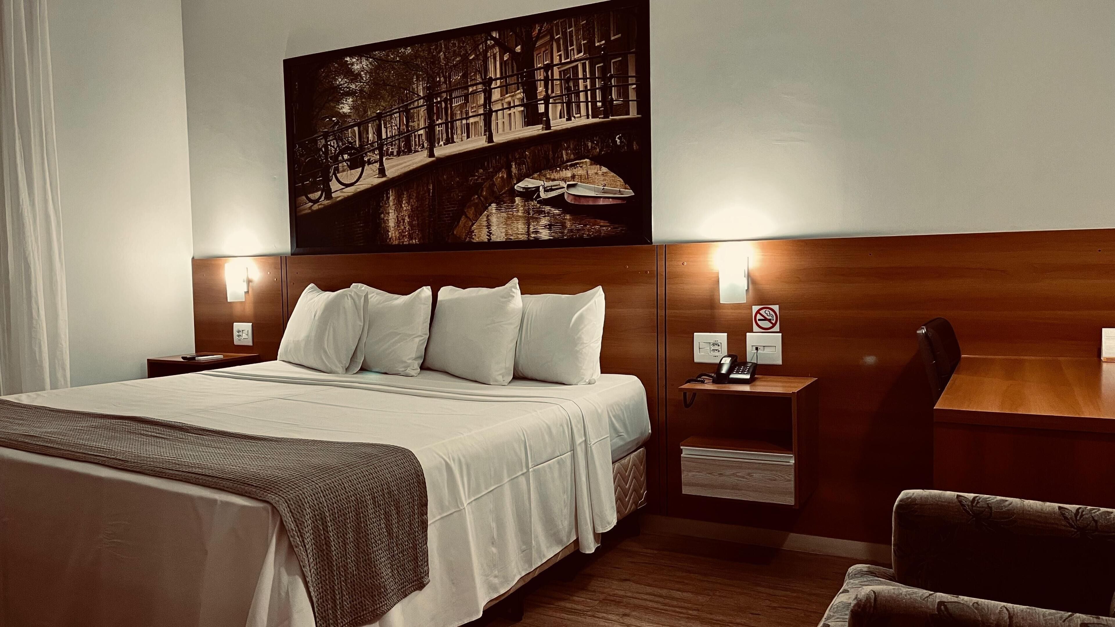 Quarto de hotel com uma cama grande, cabeceira de madeira, mesa de cabeceira com telefone e luminária fixa na parede.