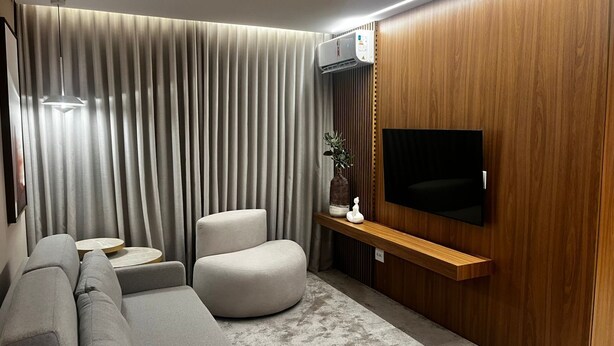 A modern living room with a grey sofa, a round coffee table, a cushioned armchair, a wooden TV stand, and a large flat-screen TV. - Hotel Amsterdam Montes Claros (Montes Claros)