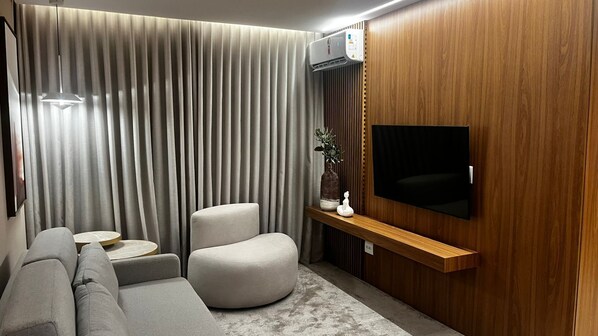 A modern living room with a grey sofa, a round coffee table, a cushioned armchair, a wooden TV stand, and a large flat-screen TV. - Hotel Amsterdam Montes Claros (Montes Claros)