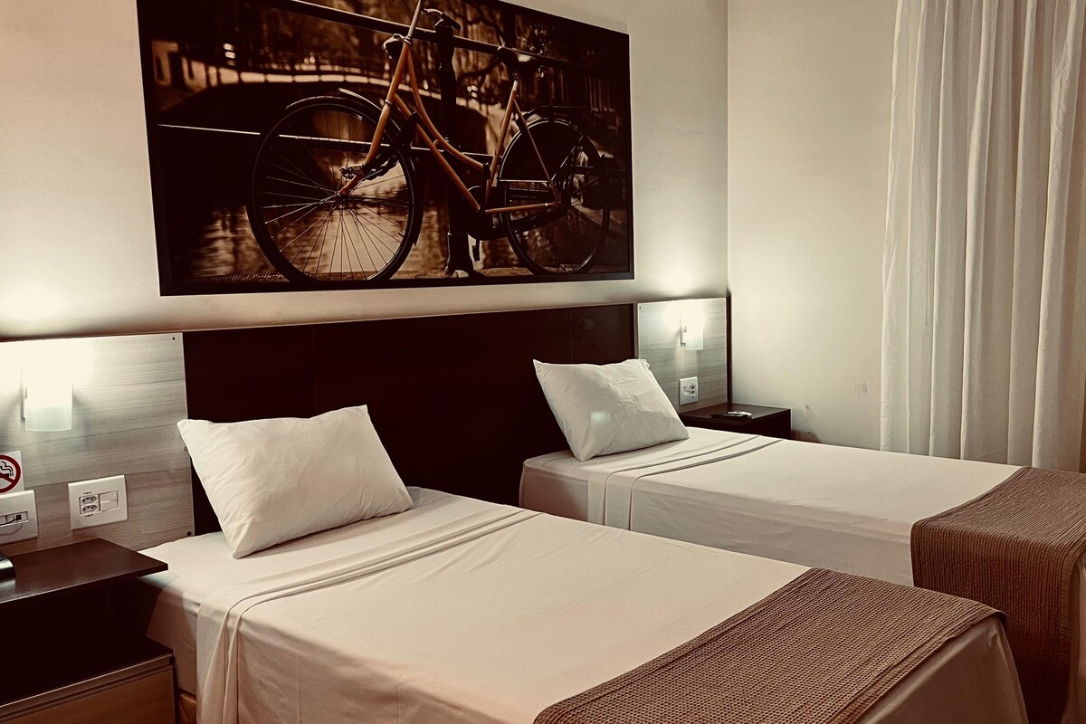 A hotel room with two single beds, a nightstand, a lamp, a painting of a bicycle, and a window with curtains.