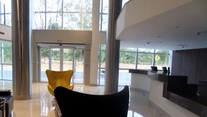A modern reception area with a glass door leading outside, a reception desk, and a yellow chair. - Hotel Amsterdam Montes Claros (Montes Claros)