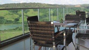 Una terraza con barandillas de vidrio, sillas de mimbre y una mesa con superficie de vidrio, con vistas a un paisaje verde y una masa de agua.