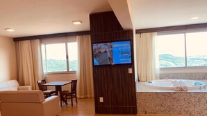 Habitación de hotel con una televisión grande, zona de comedor, sofá, bañera y vistas a las colinas.
