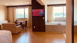 Habitación de hotel con cama, zona de estar con mesa y sillas, televisor montado en la pared y bañera con vistas al exterior.