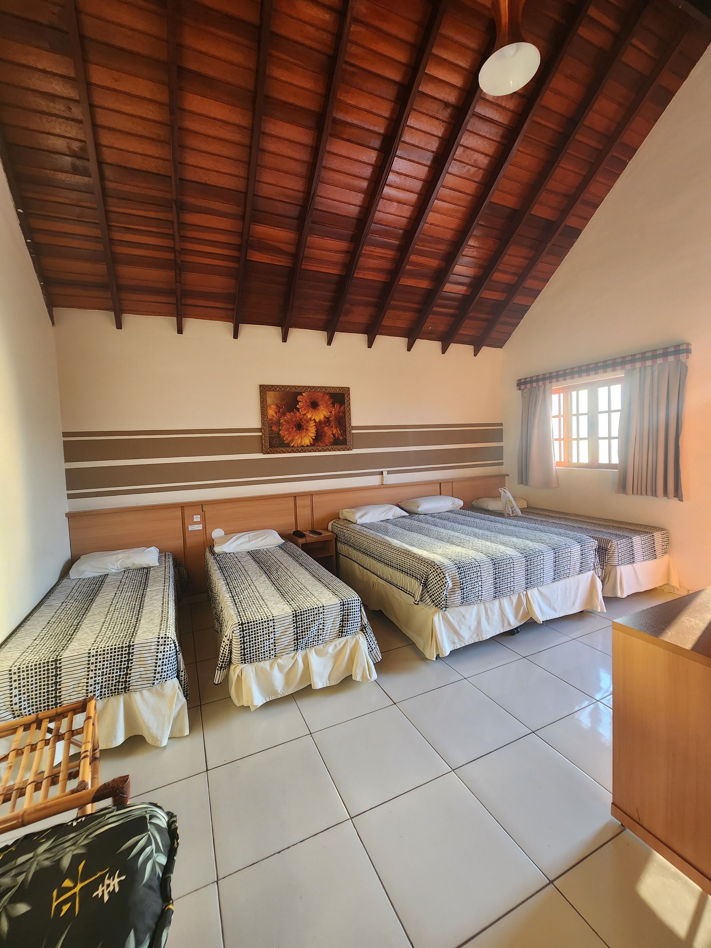 Habitación de hotel con tres camas individuales, techo de madera, un cuadro en la pared y ventana con cortinas.