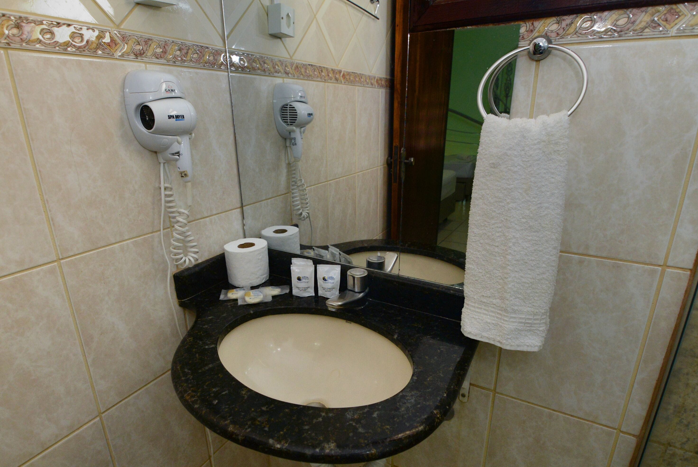 Un baño con un lavabo, dos secadores de pelo montados en la pared, una toalla colgada en un perchero y artículos de aseo sobre el mostrador.