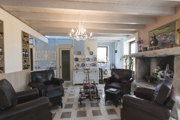 A cozy living room with leather armchairs, a wooden coffee table, and a chandelier. There is a fireplace with a stone mantle and a collection of decorative plates on the wall. - Palascia Wellness Relais (Otranto)