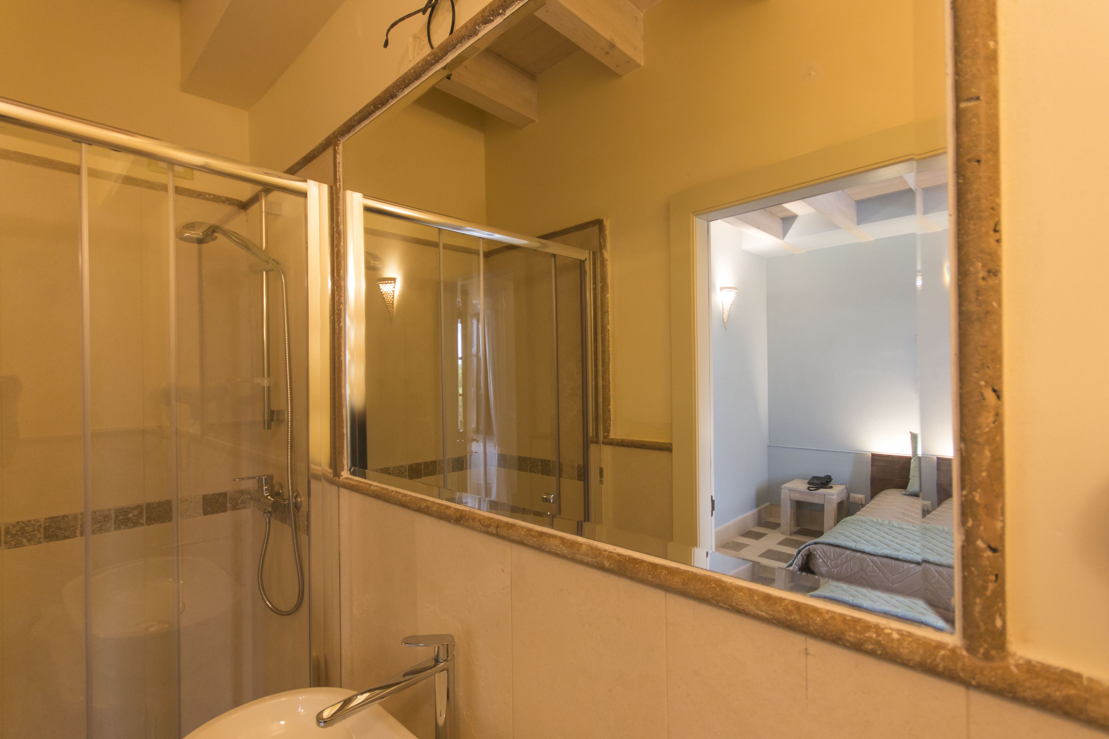 Un baño con mampara de vidrio, lavamanos y un espejo grande que refleja un dormitorio con cama y mesita de noche.