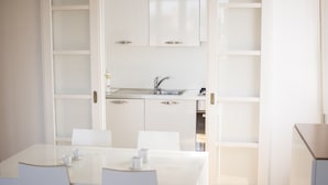 Una cocina moderna con armarios blancos, un lavamanos de acero inoxidable y una zona de comedor con una mesa de vidrio y sillas blancas.