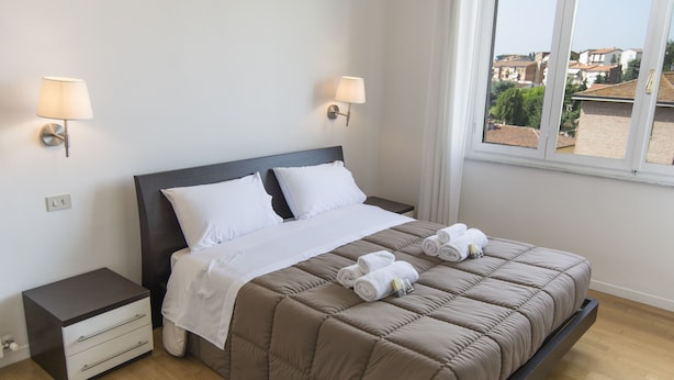Una cama bien hecha con un edredón marrón, dos almohadas blancas y toallas enrolladas sobre la cama. Hay dos mesitas de noche con lámparas, un pequeño aparador y una ventana con vistas a edificios.
