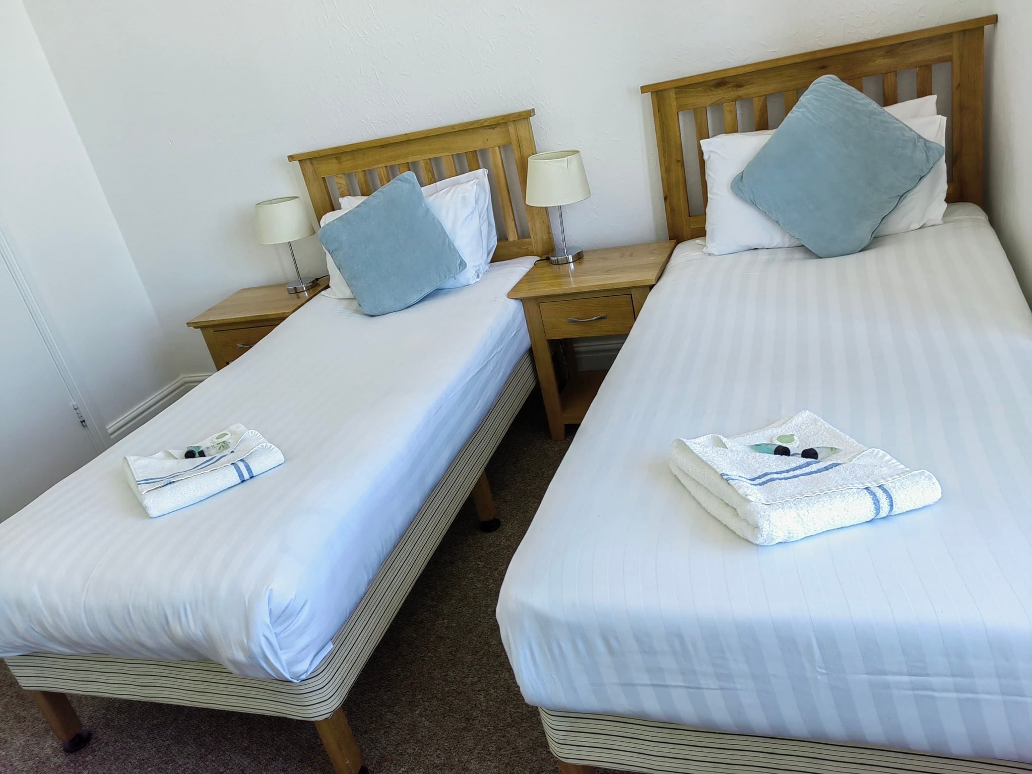 Two single beds with wooden headboards, white linens, and blue pillows. Each bed has a bedside table with a lamp and a folded towel.