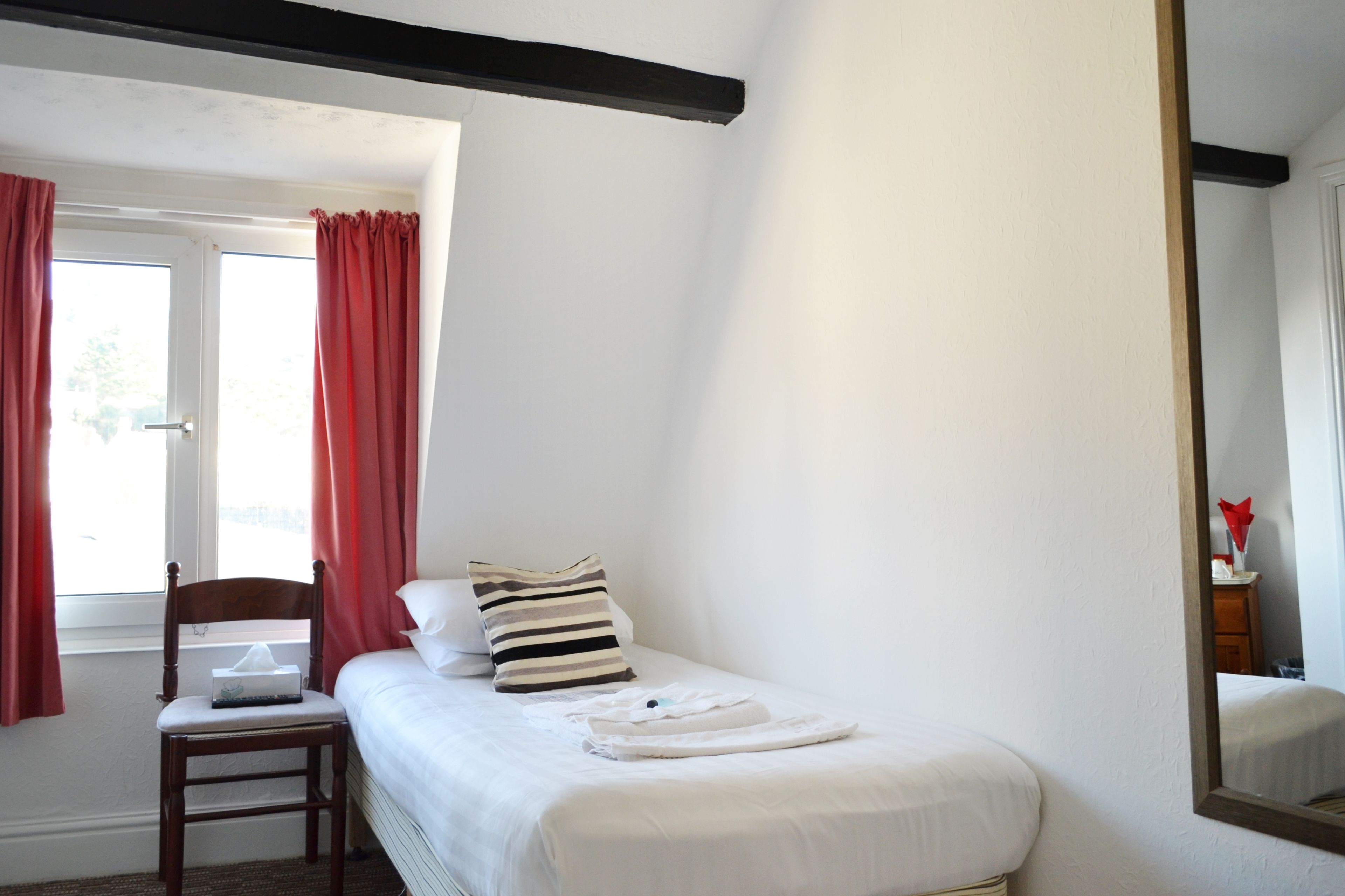 Une chambre à coucher avec un lit simple, une chaise en bois, une table de chevet et une fenêtre avec des rideaux rouges.