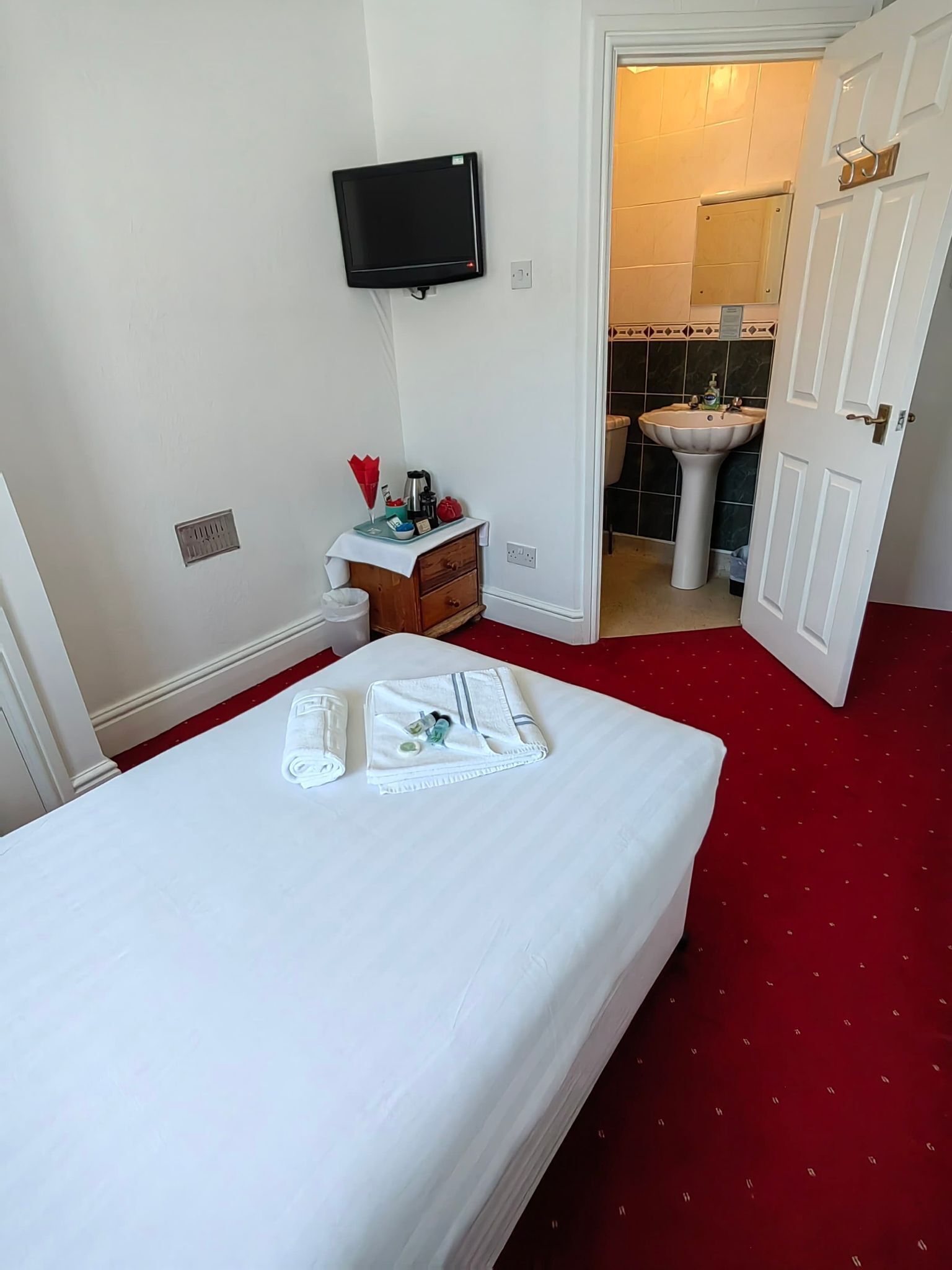 Een hotelkamer met een bed, een aan de muur bevestigde televisie, een klein tafeltje met toiletartikelen, en een badkamer zichtbaar door een open deur.
