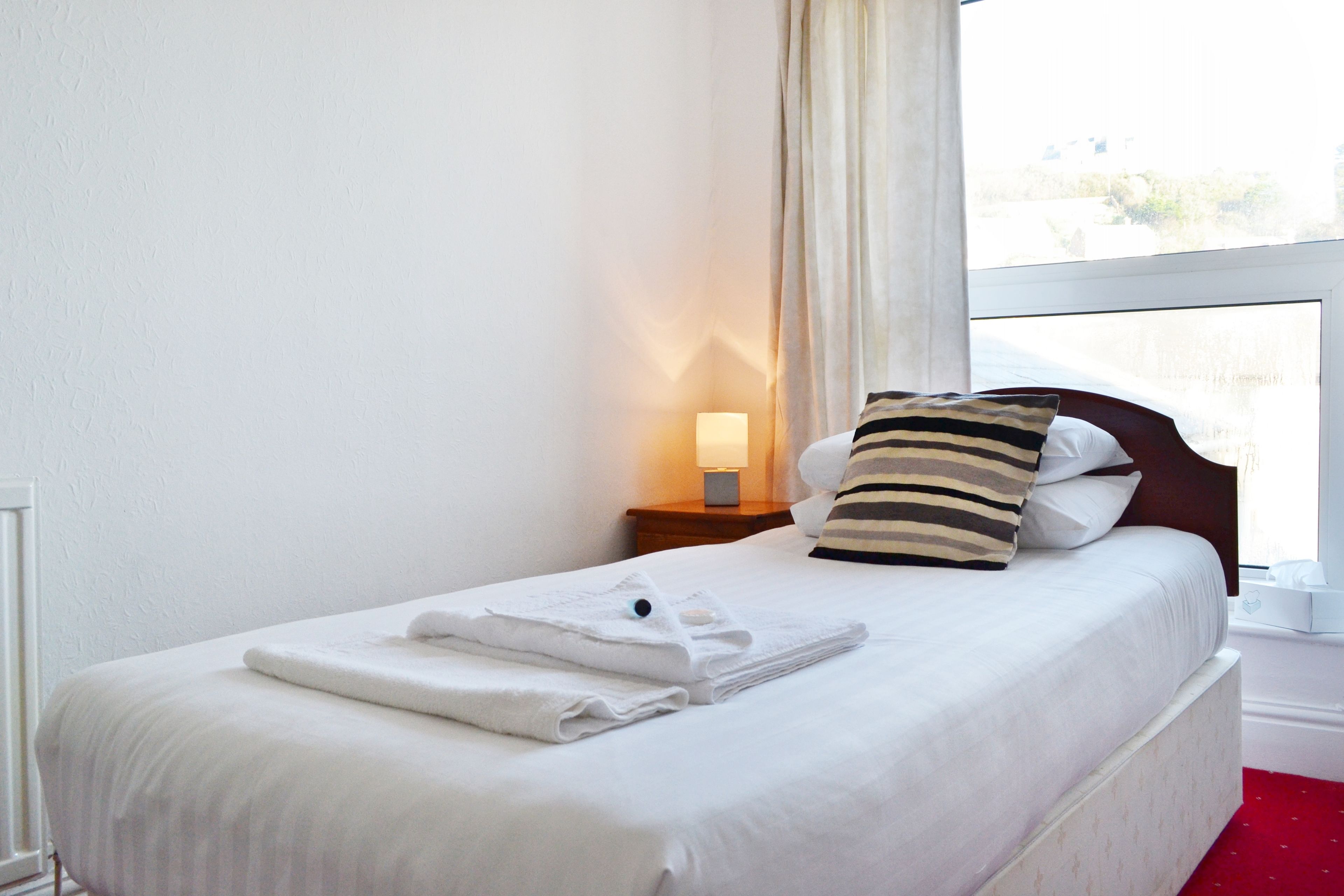A neatly made bed with white linens and a striped pillow, a bedside lamp, and a window with curtains.