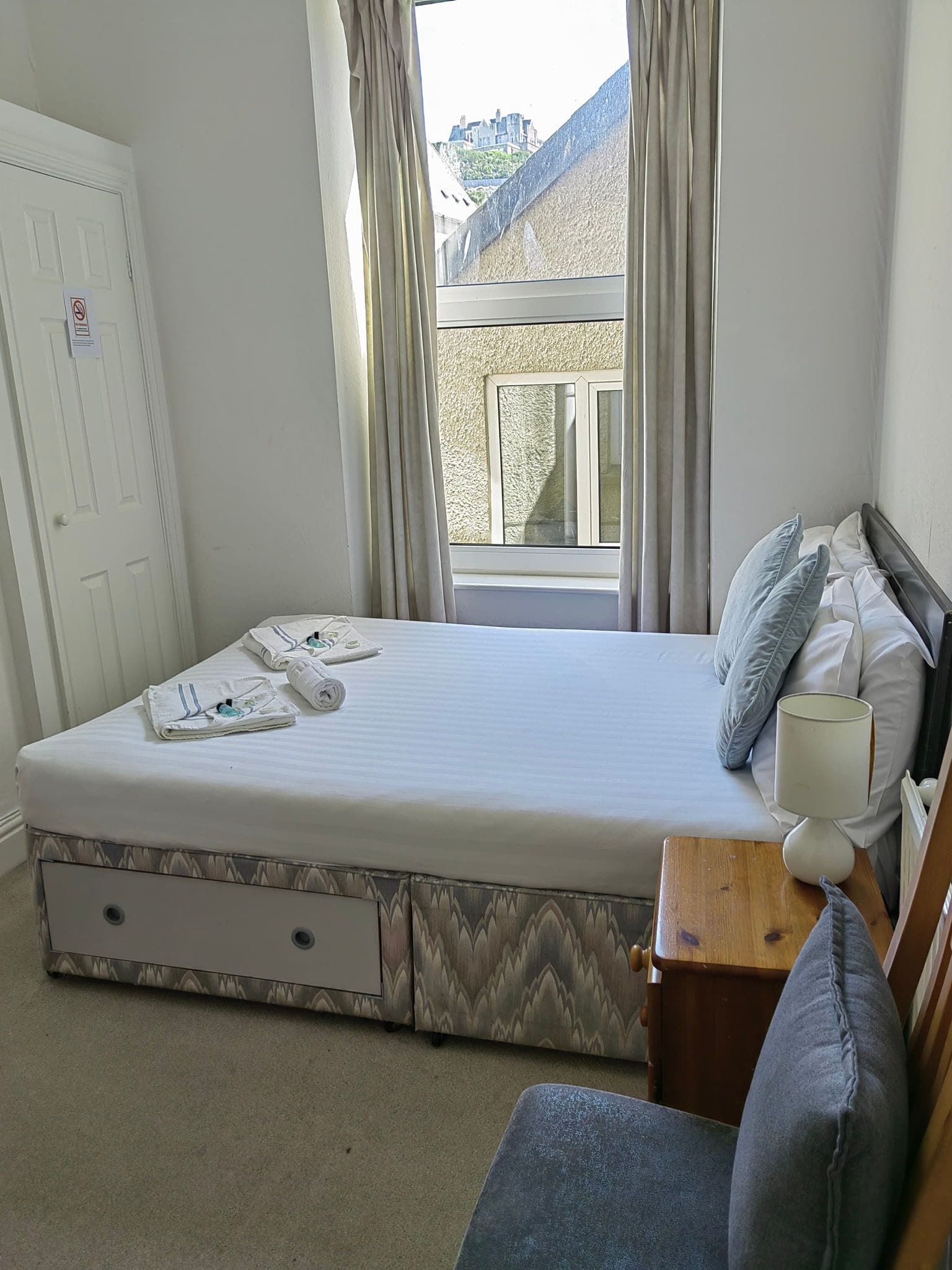 A bedroom with a bed, a bedside table with a lamp, a chair, and a window with curtains.