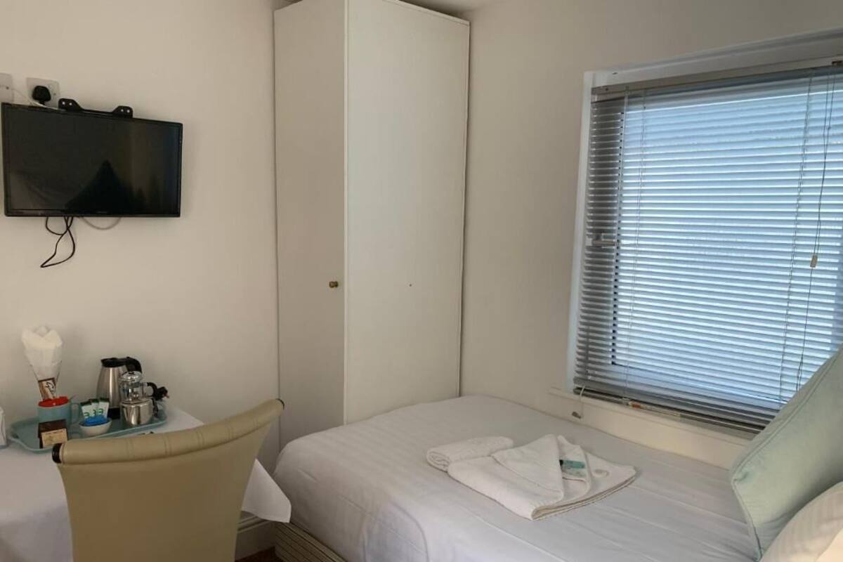 Ein Hotelzimmer mit einem Bett, einem an der Wand montierten Fernseher, einem Fenster mit Jalousien und einem kleinen Tisch mit einer Tablettladung.