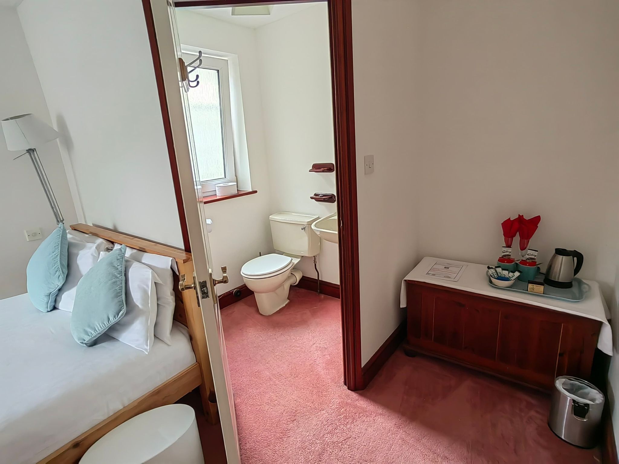 A small, compact hotel room with a bed, a bedside table, a desk, a chair, a toilet, a sink, and a small window.