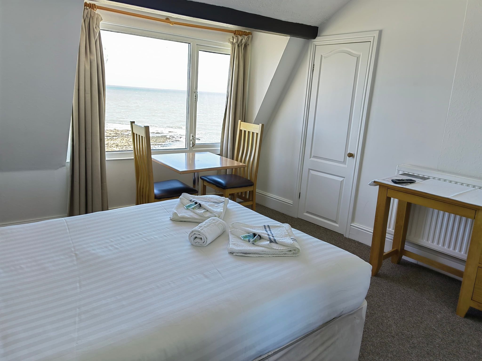 Une chambre d’hôtel équipée d’un lit, d’un bureau, d’une chaise et offrant une vue sur la mer grâce à de grandes fenêtres.