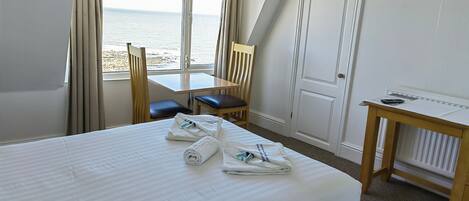 Une chambre d’hôtel équipée d’un lit, d’un bureau, d’une chaise et offrant une vue sur la mer grâce à de grandes fenêtres.