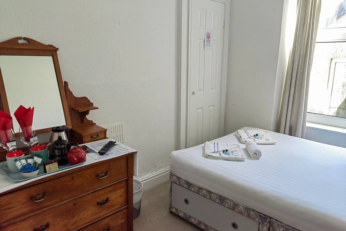 Ein Schlafzimmer mit einem Holzkapständer, einem Spiegel, einem Bett mit gemustertem Bettbezug und einem Fenster mit Vorhängen.