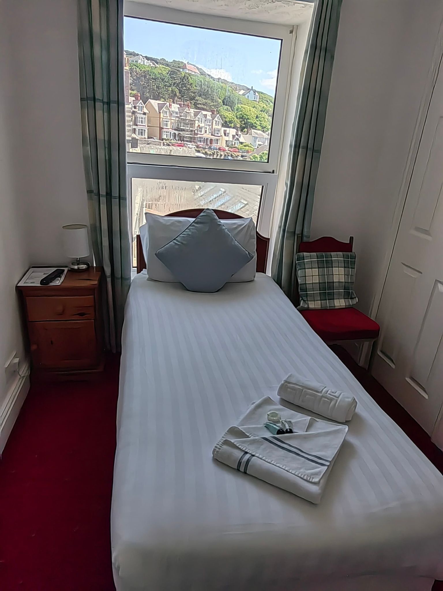 A hotel room with a bed, a nightstand, a window with a view of buildings, and a red carpet.