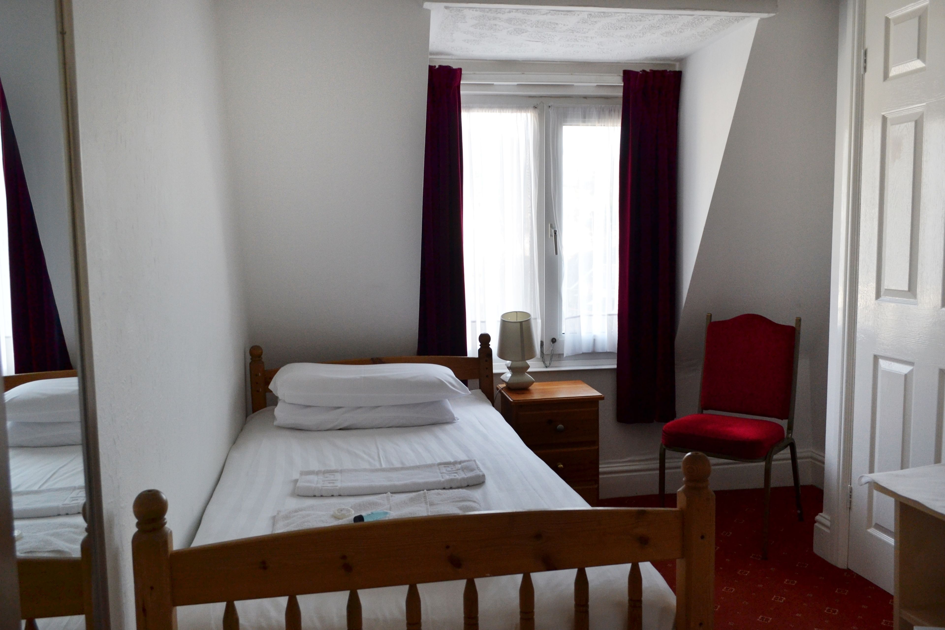Une chambre avec un lit simple, une table de chevet, une chaise et une fenêtre avec des rideaux.