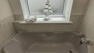 Ein Badezimmer mit einer großen Badewanne, einem Fenster mit Blick auf ein Segelboot und einem Ablagefach mit Handtüchern und Pflegeprodukten.