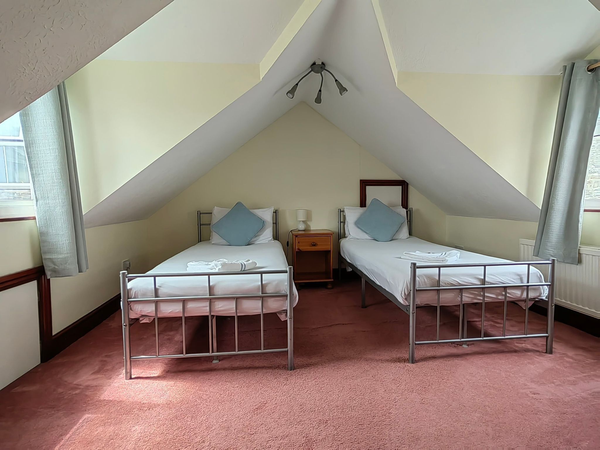 Un pequeño dormitorio en ático de forma triangular con dos camas litera metálicas, una mesita de noche y un ventilador de techo.