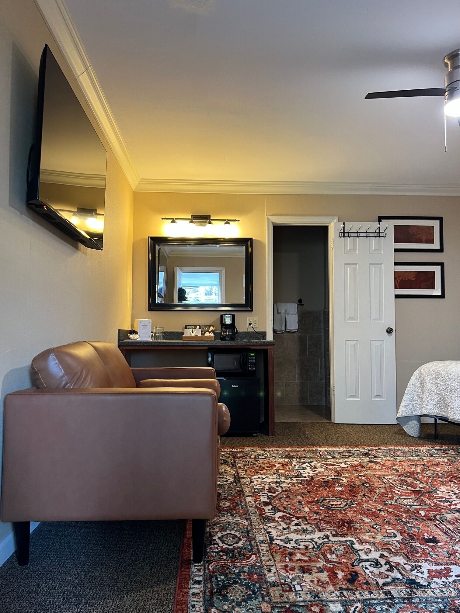 A hotel room with a sofa, a desk with a computer, a flat-screen TV, a large mirror, a door, and a patterned rug.