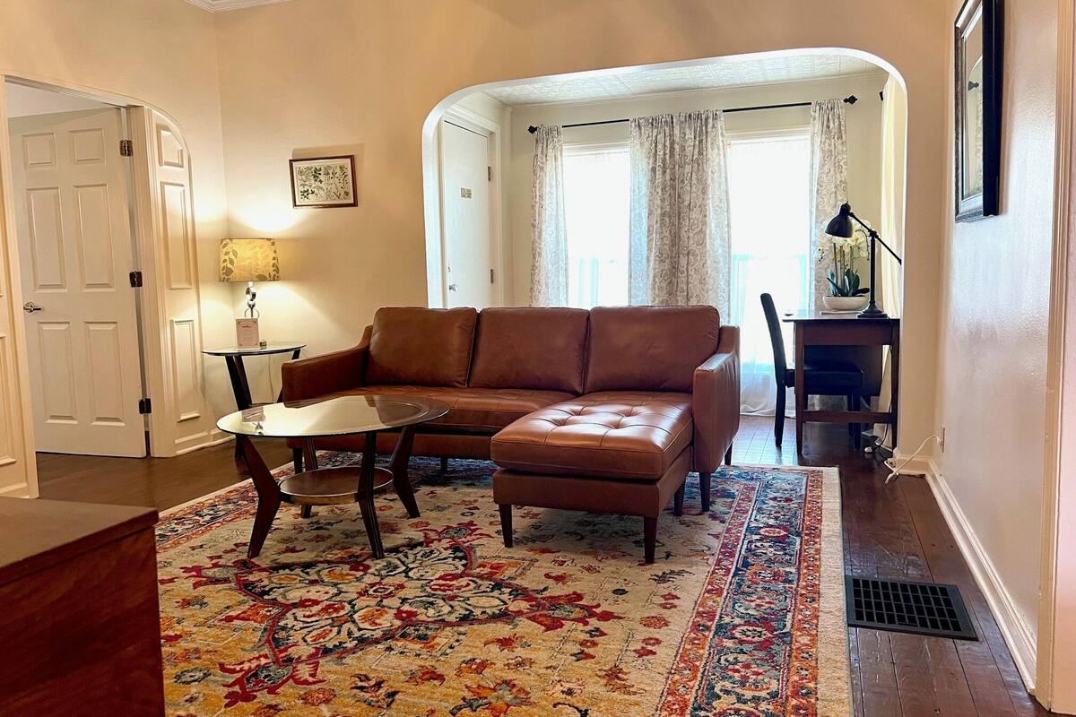 A living room with a brown leather sofa, a glass coffee table, and a patterned rug. There is a dining area with a table and chairs, and a door leading to another room.