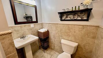 A bathroom with a sink, toilet, and a mirror with lights. There is a shelf with decorative items and a basket with towels.