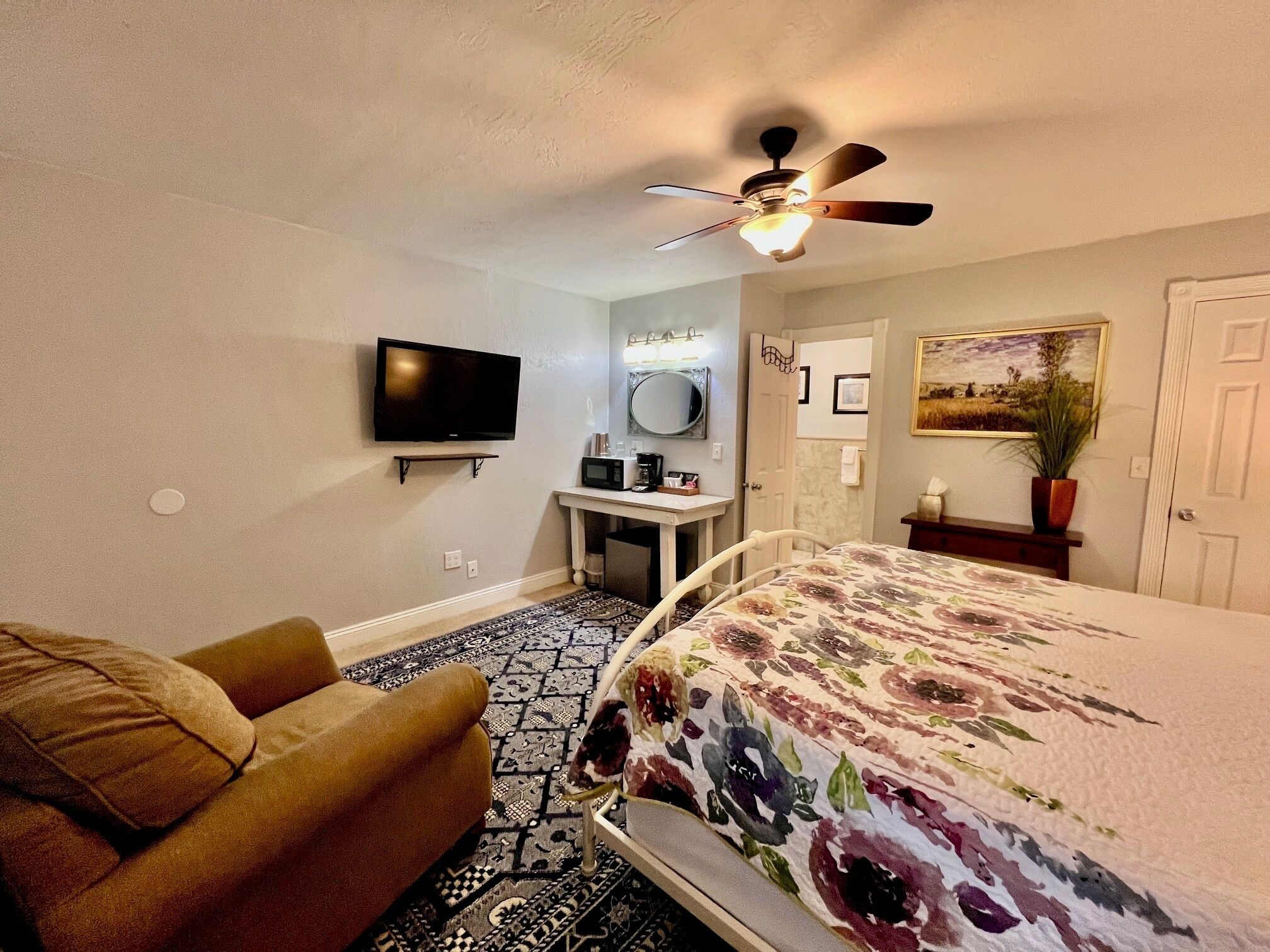 Une chambre à coucher comprenant un lit, un canapé, un ventilateur de plafond, une télévision, une petite table, un miroir et une porte.