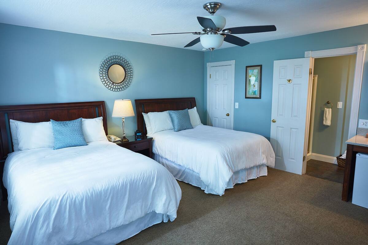 A hotel room with two beds, a ceiling fan, a mirror, and a door leading to another room.