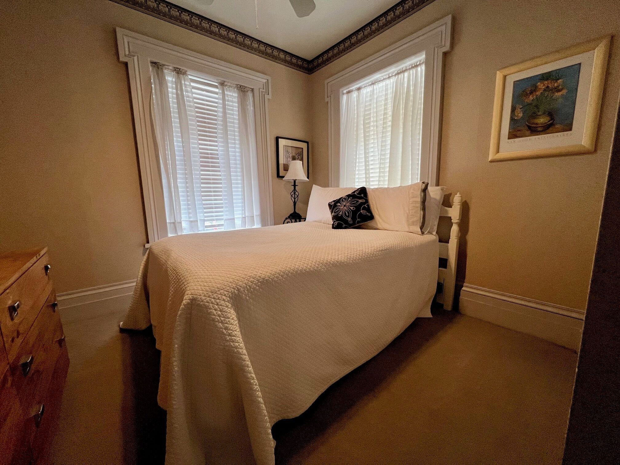 A neatly arranged bedroom with a large bed, two windows with blinds, a bedside table with a lamp, and a framed picture on the wall.