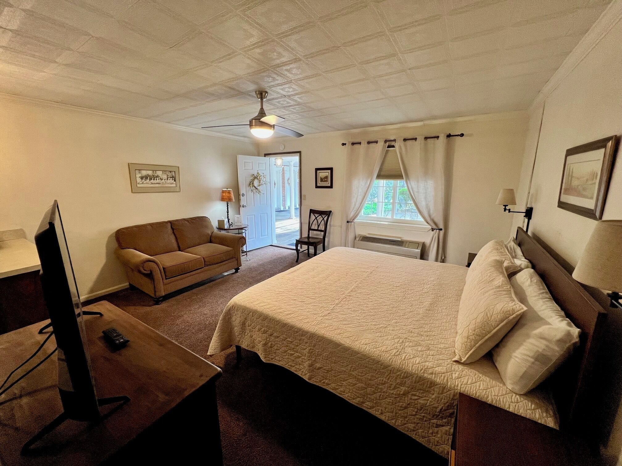 A hotel room with a bed, a desk with a computer, a sofa, a chair, a ceiling fan, a window with curtains, and a door.
