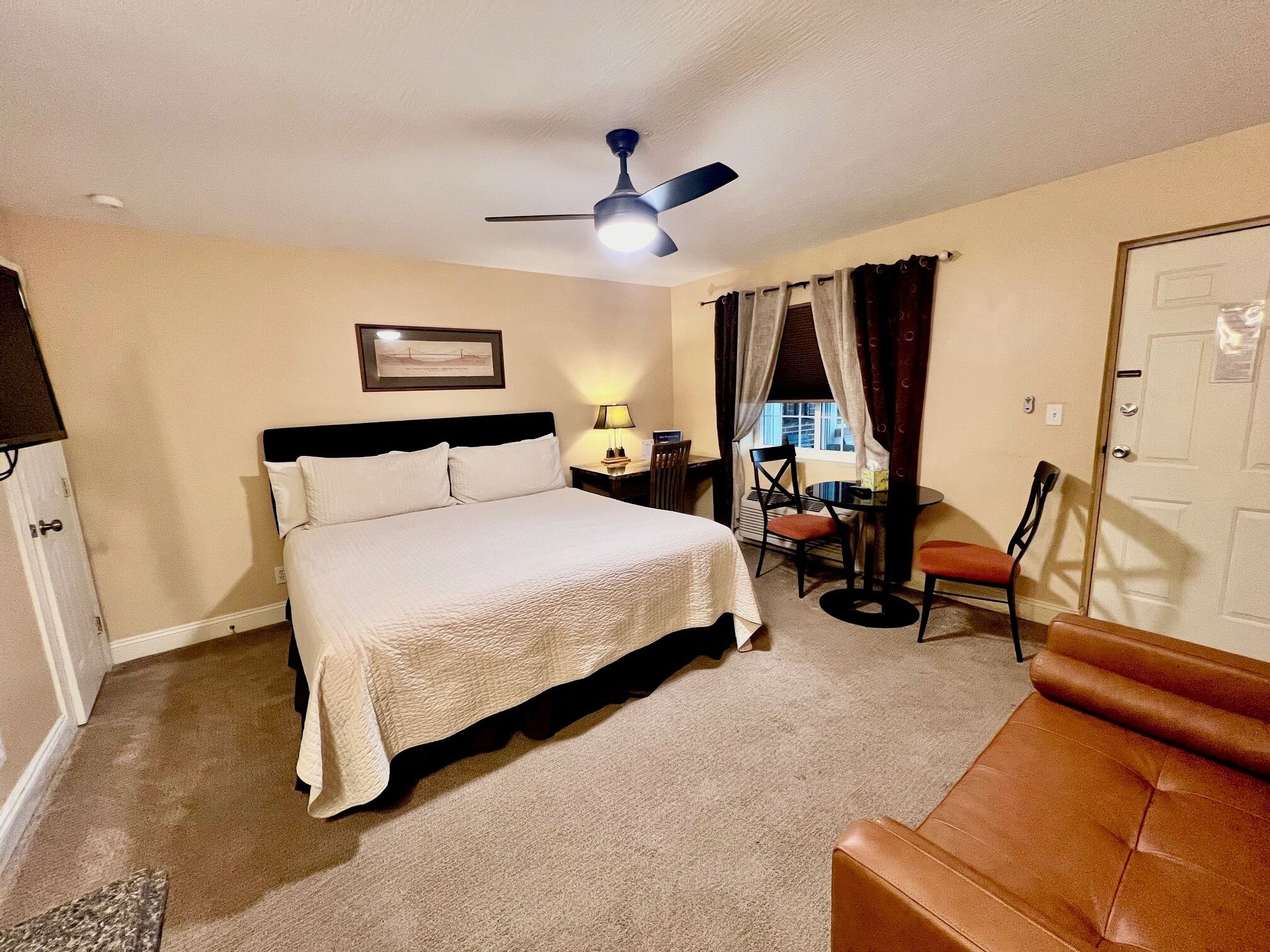 A hotel room with a bed, a desk with a chair, a ceiling fan, and a door.