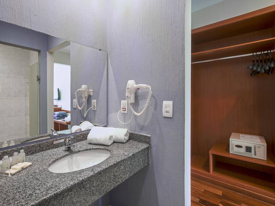 Un baño con lavamanos de granito, un espejo, un secador de pelo y un estante de madera con artículos de aseo.