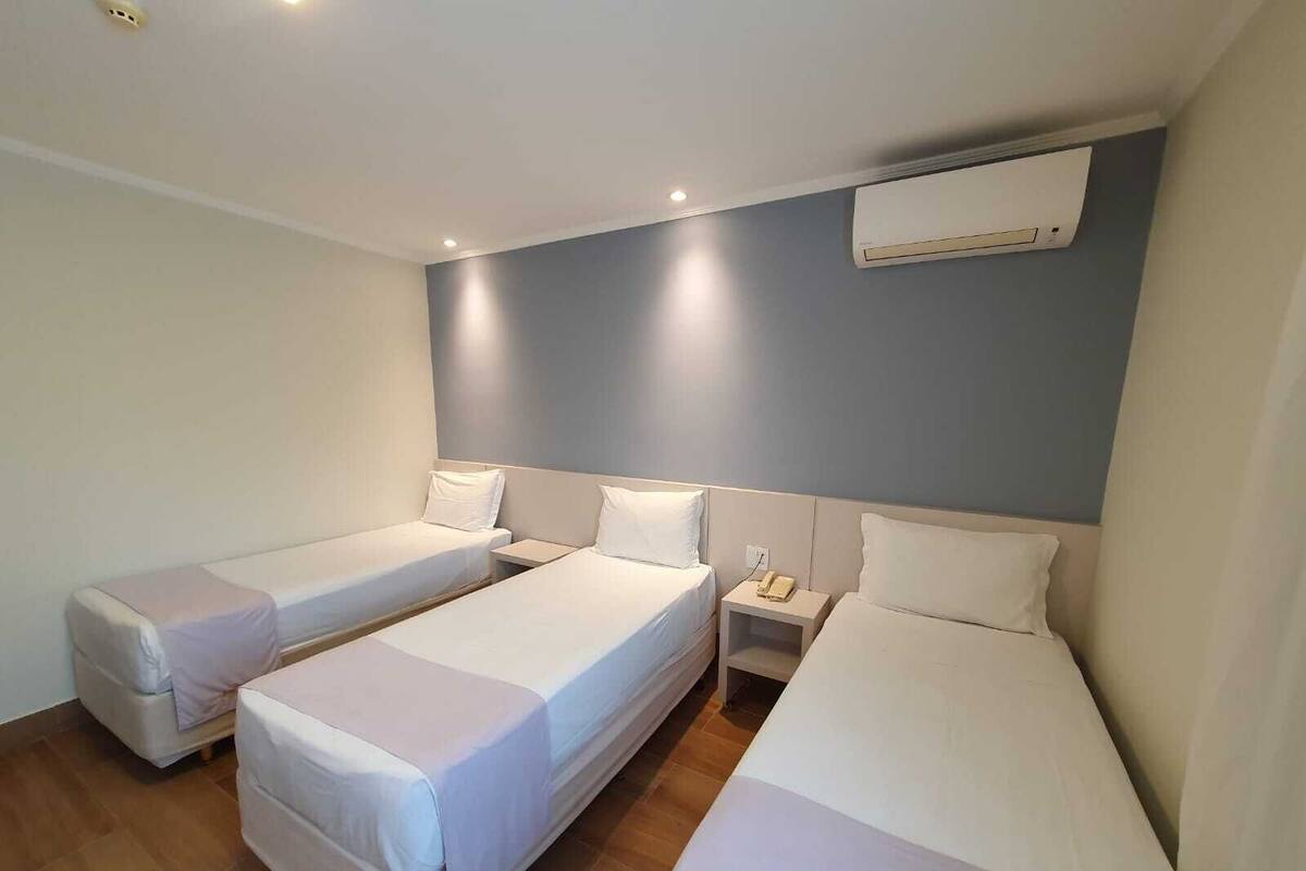 Habitación de hotel con tres camas individuales, una mesita de noche con lámpara y un equipo de aire acondicionado en la pared.