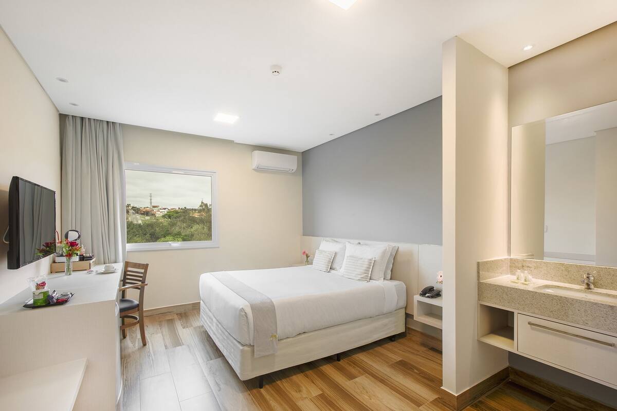 Una habitación de hotel moderna con cama, escritorio, silla, ventana grande, baño con espejo y equipo de aire acondicionado.