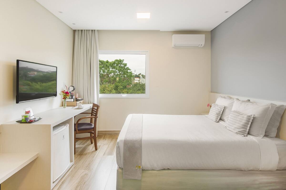 Habitación de hotel con cama, escritorio, silla, televisor y ventana con vista a árboles.
